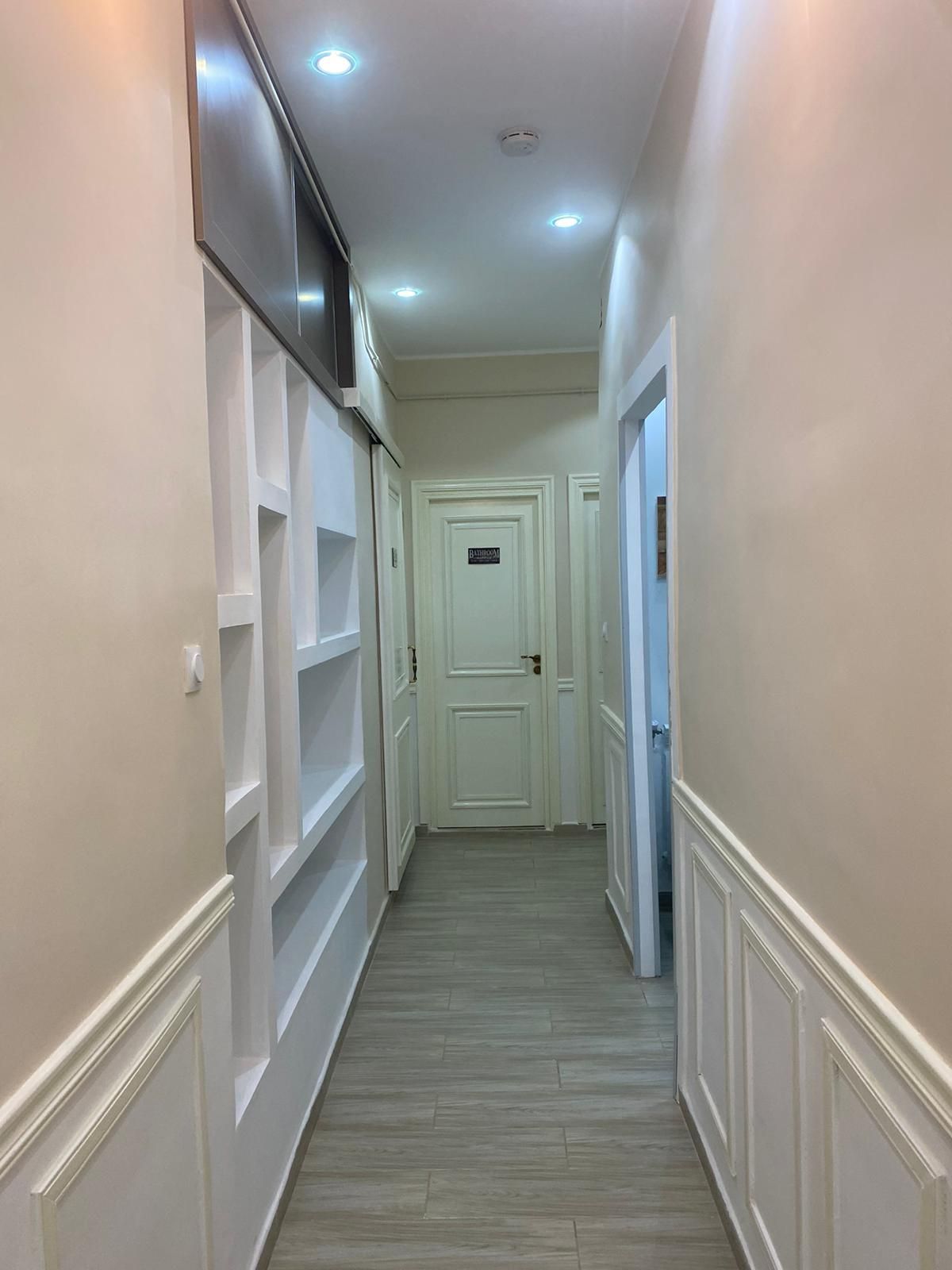 A narrow hallway with white walls, a door at the end, and a flat-screen TV mounted on the wall.