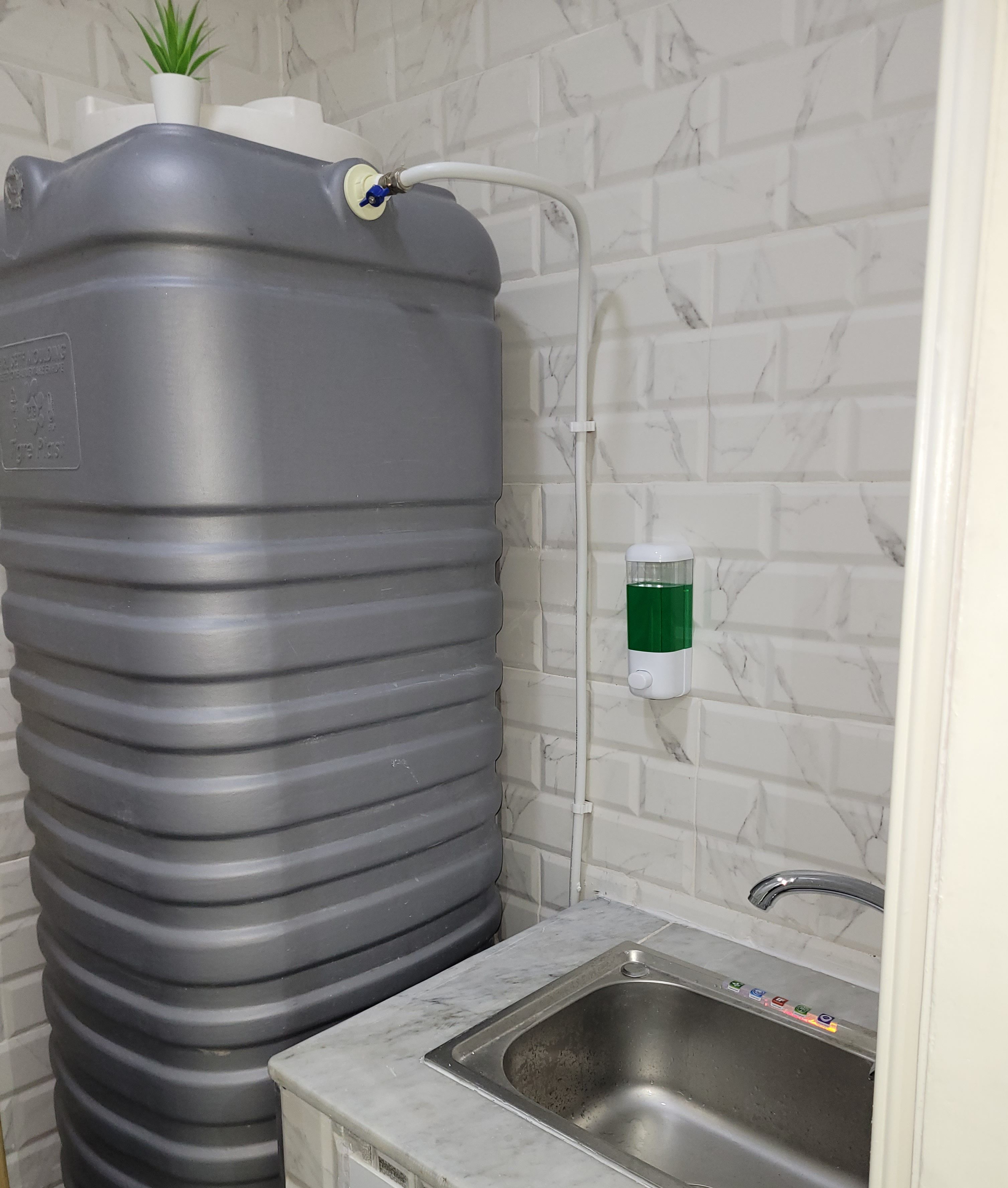 A bathroom with a large grey water tank, a green soap dispenser, and a stainless steel sink.