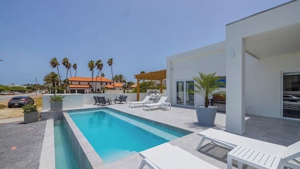 A modern outdoor swimming pool with lounge chairs, a pergola, and a view of palm trees and buildings in the background.