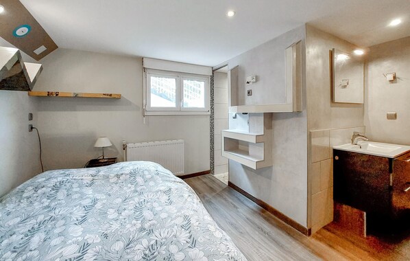 Un dormitorio con cama, mesita de noche, lámpara, ventana, radiador, armario empotrado y baño con lavabo y espejo.