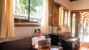 Eine Person sitzt auf einem Sofa mit einem Laptop, einem Holztisch mit einer Vase und Blick auf Grünfläche im Freien.