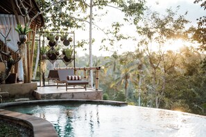 An infinity pool with a wooden deck and lounge chairs, surrounded by lush greenery and a bamboo structure.