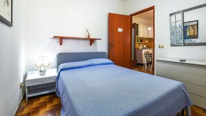 Une chambre à coucher avec un lit, une table de chevet et une petite étagère. Une porte donne accès à une petite cuisine.