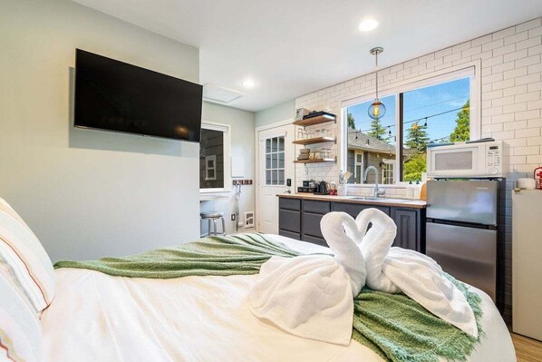 A modern hotel room with a bed, a flat-screen TV, a kitchenette, and a large window.