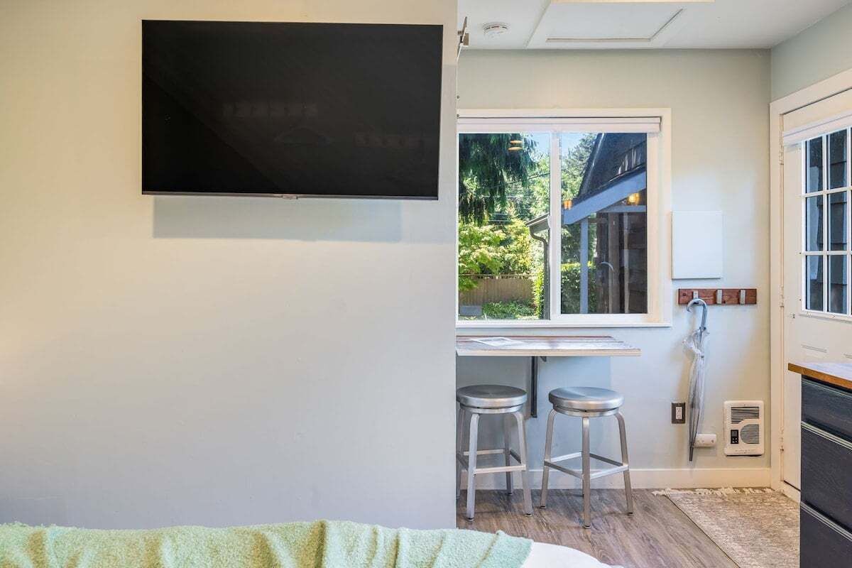 A modern room with a large flat-screen TV mounted on the wall, a window with a view of greenery, and two metal stools.