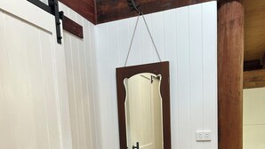 Een kamer met witte paneelwanden, een houten vloer, een deur met een zwarte hendel, een spiegel met houten lijst en een klein houten tafeltje met een plant.