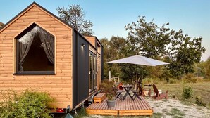 Une cabane en bois avec une grande fenêtre, une terrasse dotée de mobilier d’extérieur et un arbre en arrière-plan.