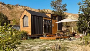 Une petite cabane en bois avec un toit noir, de grandes fenêtres et un patio aménagé avec du mobilier d’extérieur.