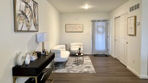 A hallway with a dark wood floor, white walls, a black console table with decorative items, a white armchair, a small round table with a lamp, and a door with a curtain. - Cozy Front Lake Home in Newtown St. Charles (Saint Charles)