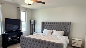 A bedroom with a tufted headboard, a flat-screen TV on a stand, a bedside table, and a fluffy white rug. - Cozy Front Lake Home in Newtown St. Charles (Saint Charles)