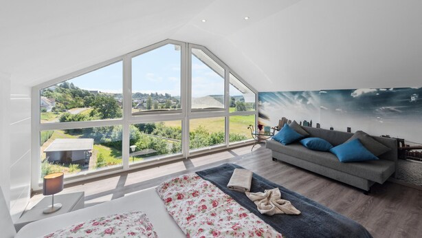 Ein geräumiges Zimmer mit einem großen Oberlicht, einem grauen Sofa mit blauen Kissen, einer blumigen Tagesdecke und einem Stadtpanorama-Wandbild.