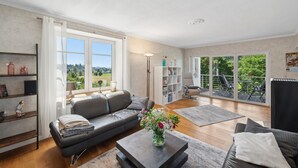 Ein geräumiges Wohnzimmer mit einer Couch, einem Couchtisch und einem Bücherregal. Ein großes Fenster bietet einen Blick ins Freie.