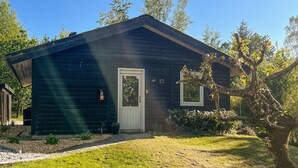 Une petite cabane en bois sombre, avec une porte blanche et deux fenêtres, entourée d’arbres et d’une cour gazonnée.