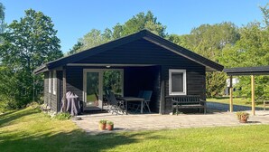 Une petite cabane en bois sombre, dotée d’un patio couvert, d’une porte-fenêtre coulissante et d’un petit espace extérieur aménagé avec des chaises et une table.