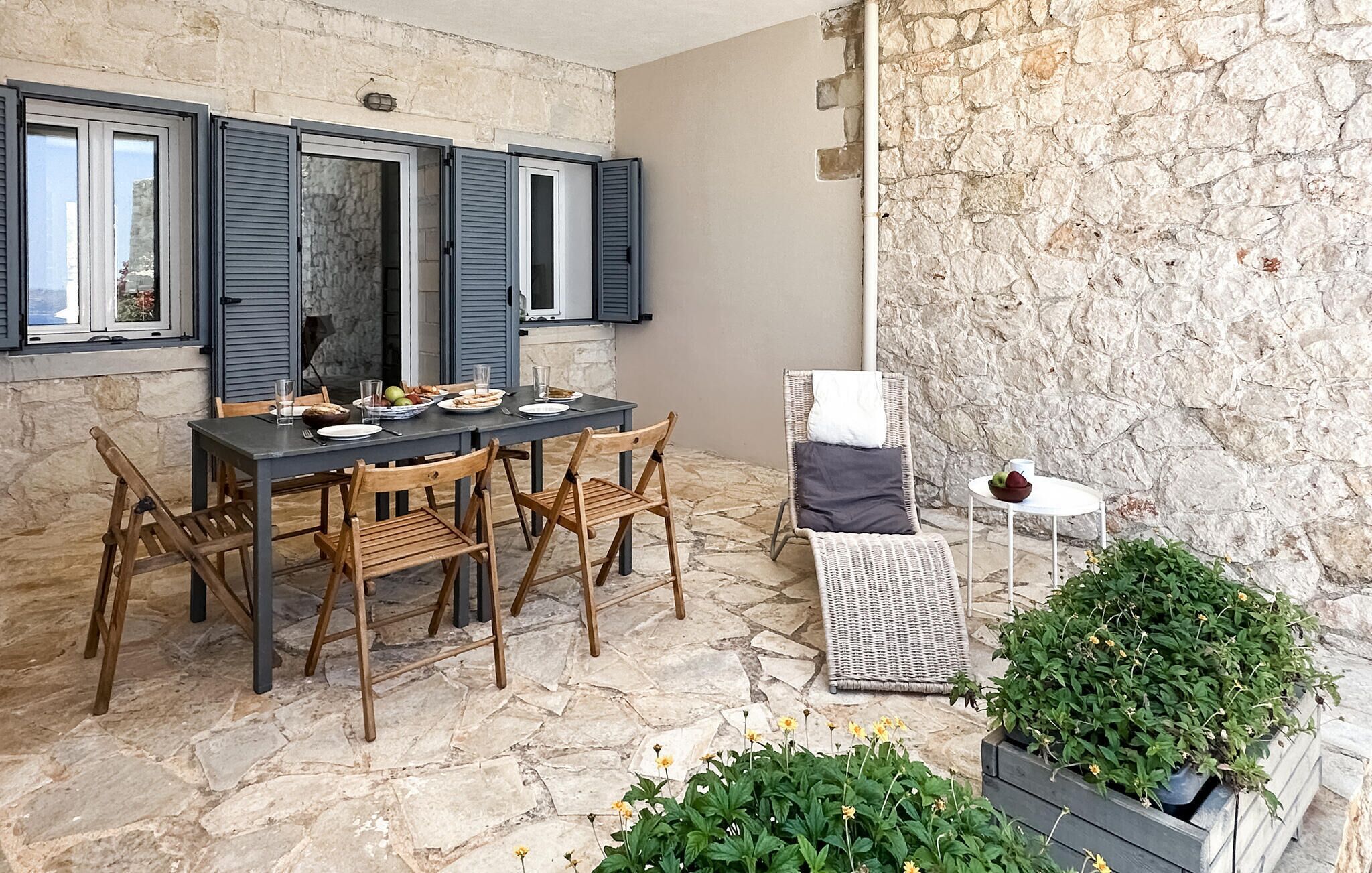 A stone-floored patio with a table set for two, wooden chairs, a wicker chair, and a small round table with a bowl.