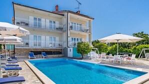 Un espace piscine doté de chaises longues et de parasols, une piscine dont le fond est recouvert de pierres, et un bâtiment avec des balcons.