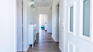 Un couloir doté d’un plancher en bois, de murs blancs et d’une porte donnant sur une chambre à coucher avec un lit et des rideaux.