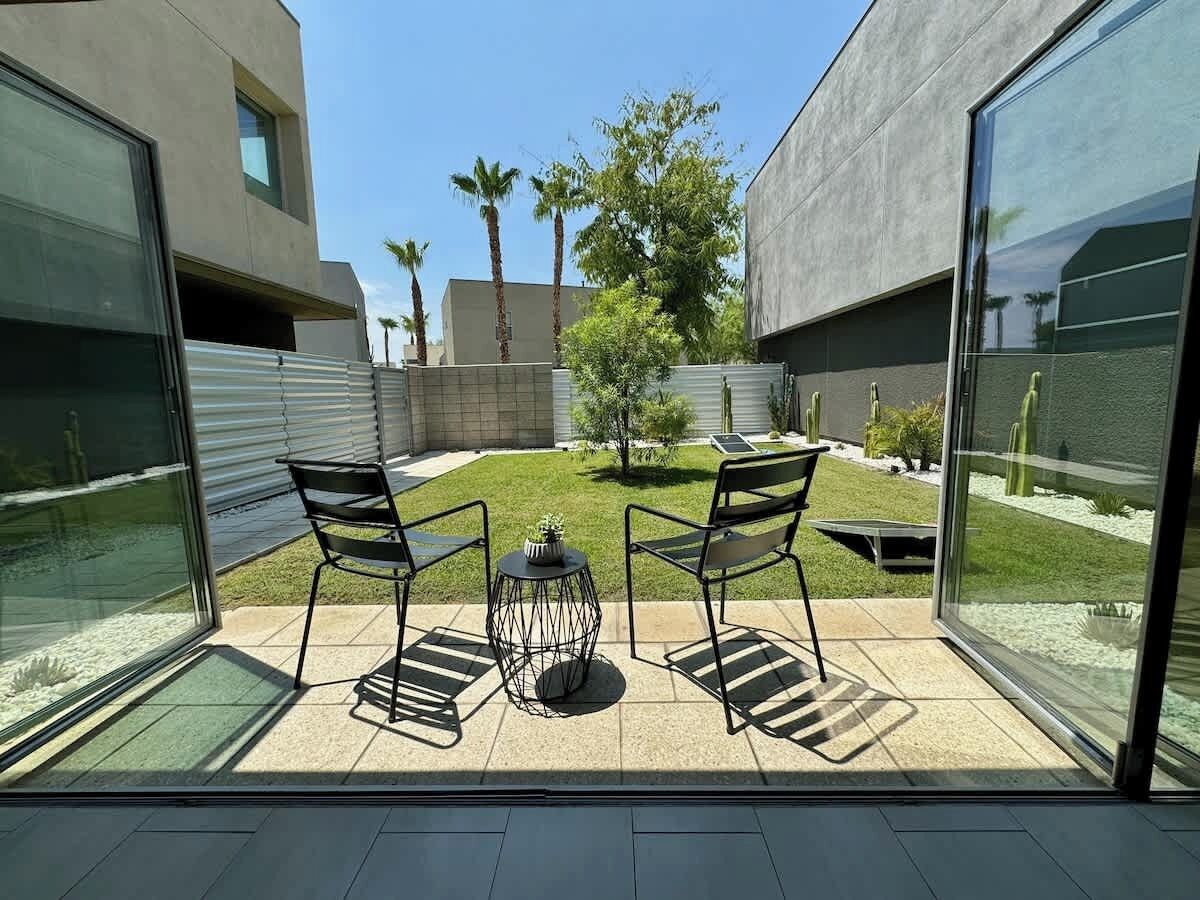 A modern patio with two chairs and a small table, a glass door leading to a garden, and a building with a concrete facade.