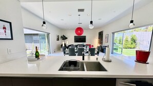 A modern kitchen with a central island, a dining area with chairs, and a large window. - New! Audrey's Luxury Retreat - Walk Downtown (Palm Springs)