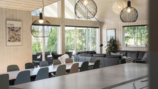 Een moderne eetruimte met een lange tafel, stoelen en een bank. Grote ramen zorgen voor natuurlijk licht en uitzicht op groen buiten.