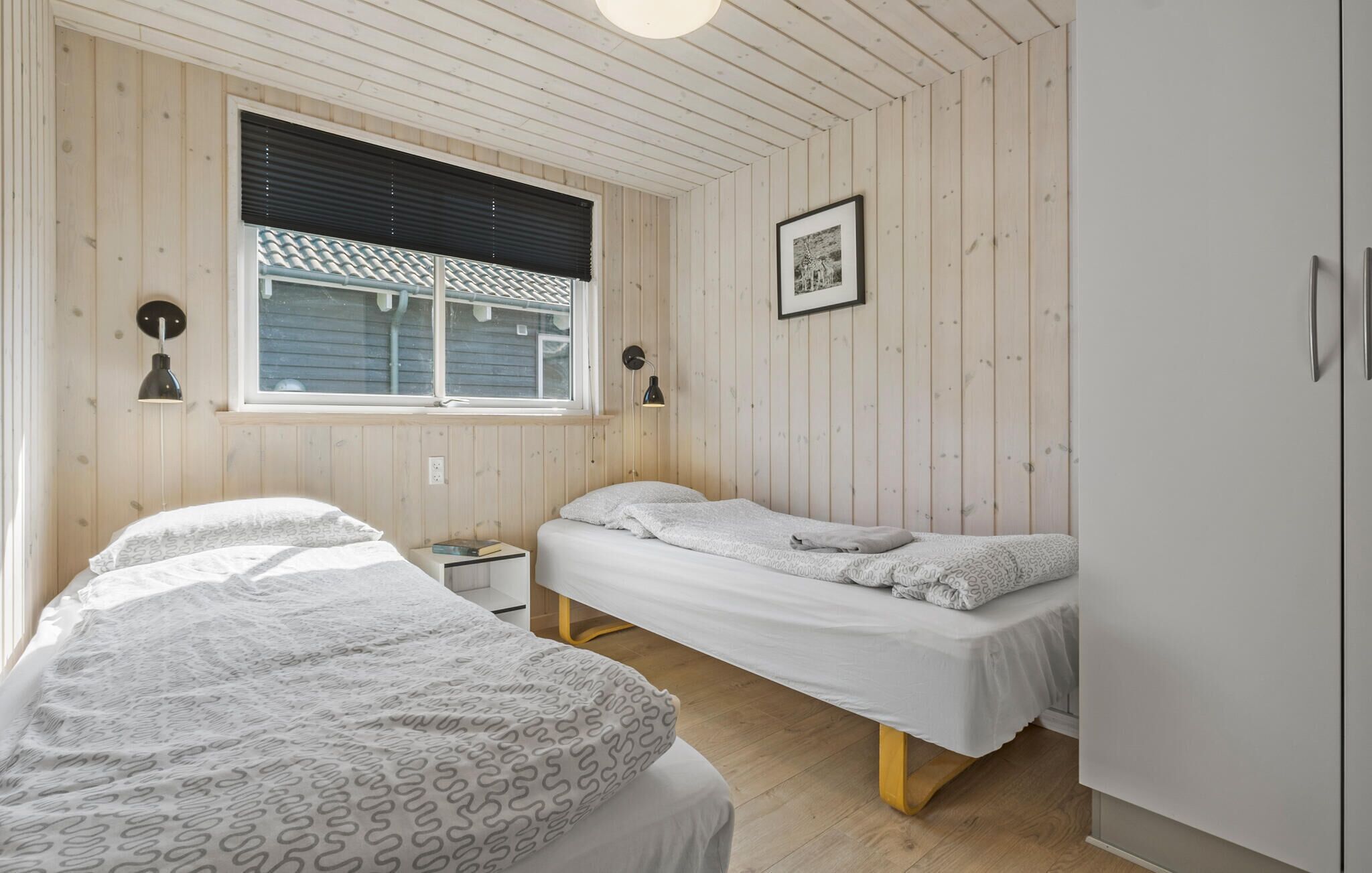 A room with two beds, wooden walls, a window with blinds, a framed picture, and a wardrobe.