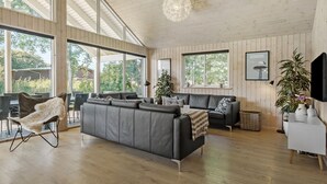 Een ruime woonkamer met een grijze bank, een eetgedeelte met een tafel en stoelen, grote ramen met uitzicht op groen, en een moderne kroonluchter.