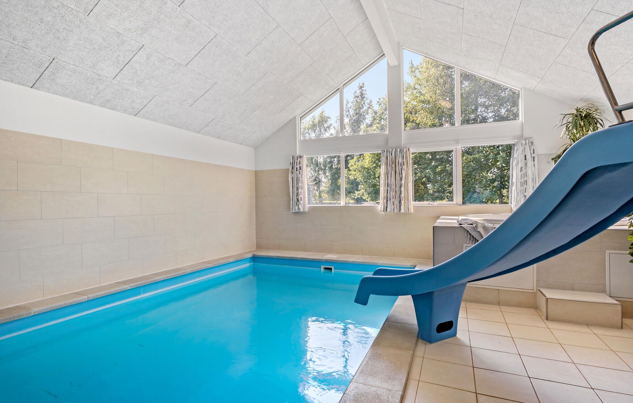 An indoor swimming pool with a blue slide, large windows, and a view of trees outside.
