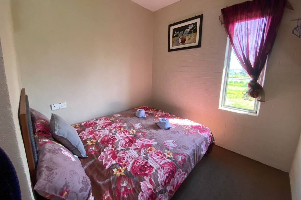 Ein kleines Zimmer mit einem Einzelbett, einer Bettdecke mit Blumenmuster, einem Fenster mit Vorhang, einem Wandbild und einer Steckdose.