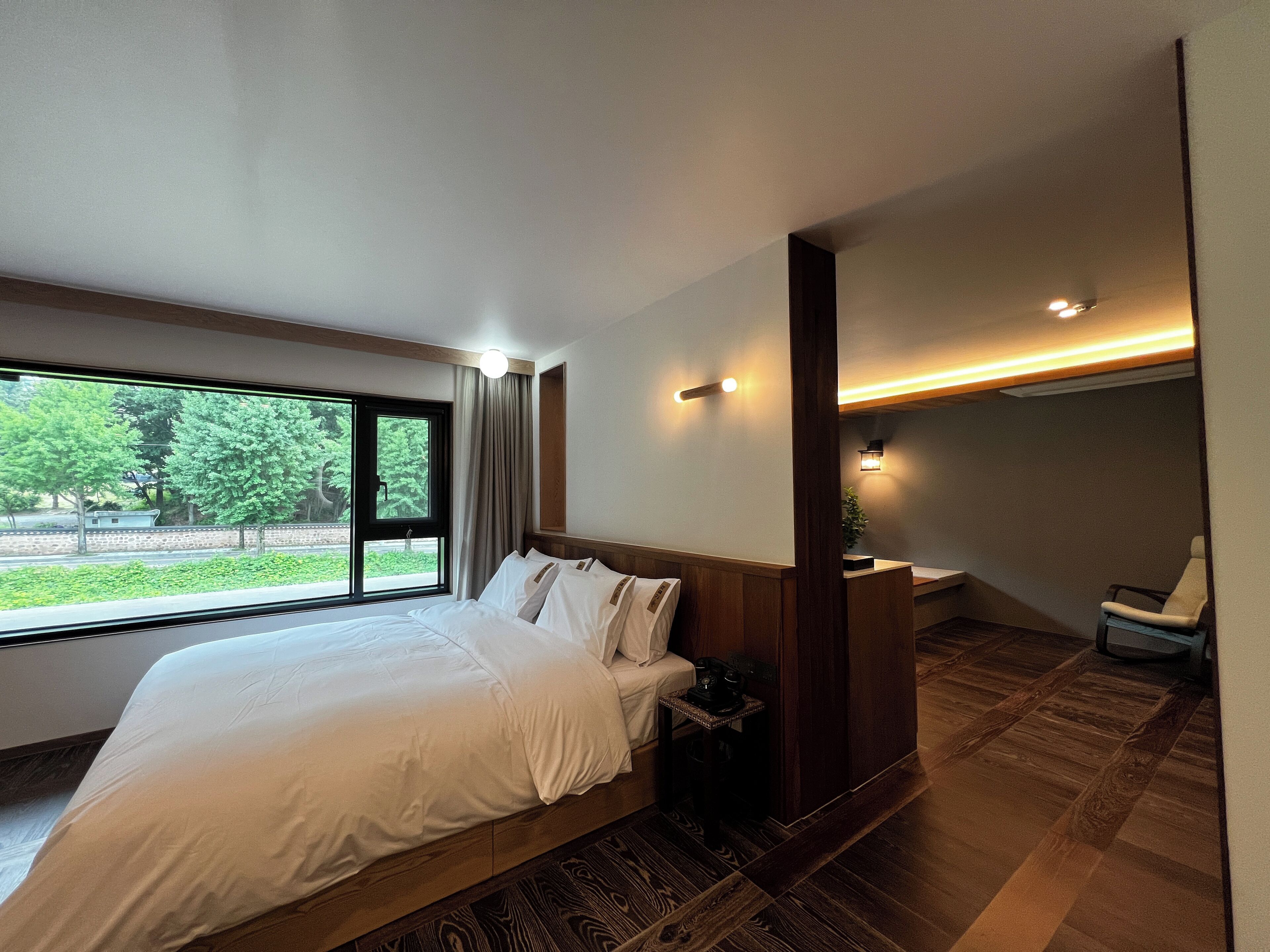 A modern hotel room with a large bed, a small desk, a chair, and a view of greenery outside.