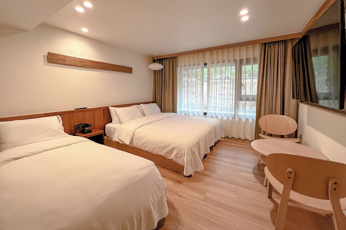 Ein Hotelzimmer mit zwei Betten, einem Holfnachtkästchen, einem Sessel und einem großen Fenster mit Vorhängen.