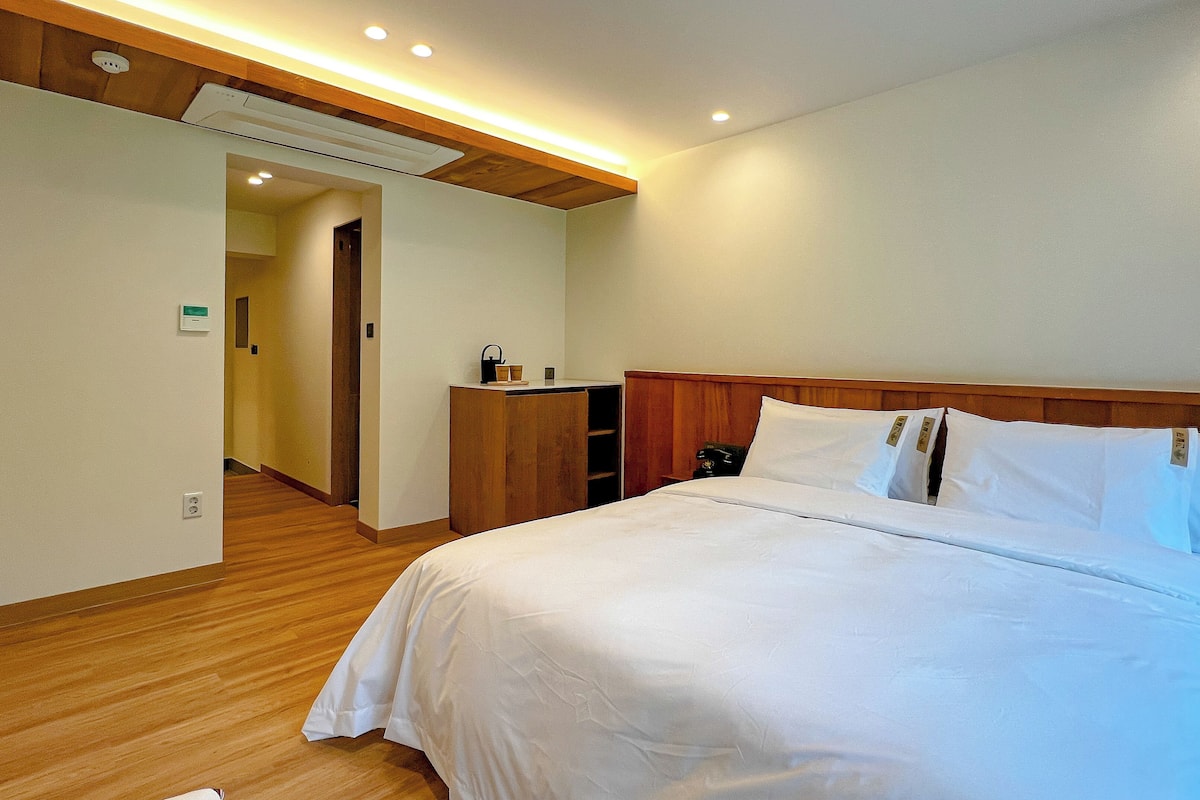 Ein Hotelzimmer mit einem großen Bett, einem Holzkopfende und Holzboden. Es gibt eine kleine Holzkommode und einen Nachttisch mit einer Lampe.