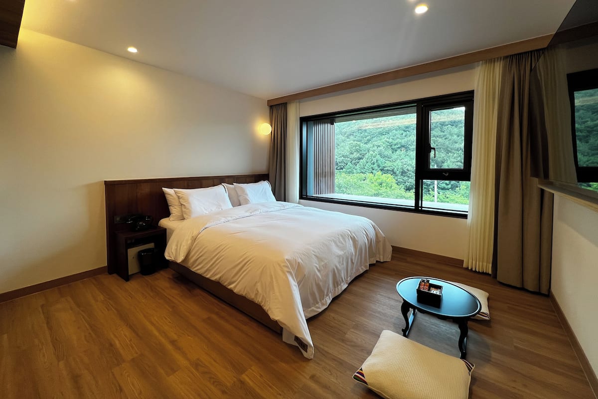 Ein Hotelzimmer mit einem großen Bett, einem kleinen Tisch und einem Fenster mit Blick auf Grünflächen.