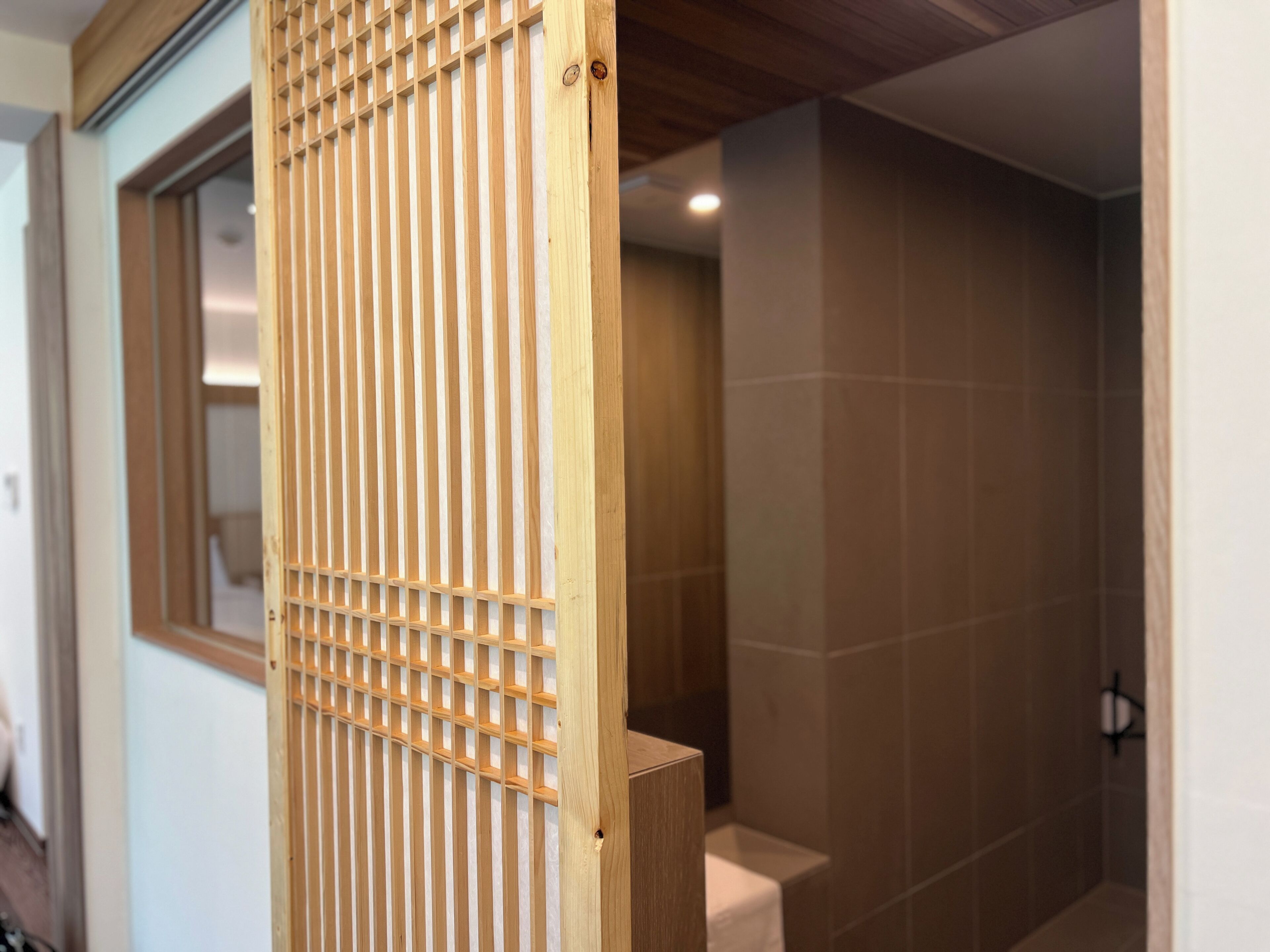 Una mampara de madera con listones verticales en un baño, una pared con azulejos y una ventana.