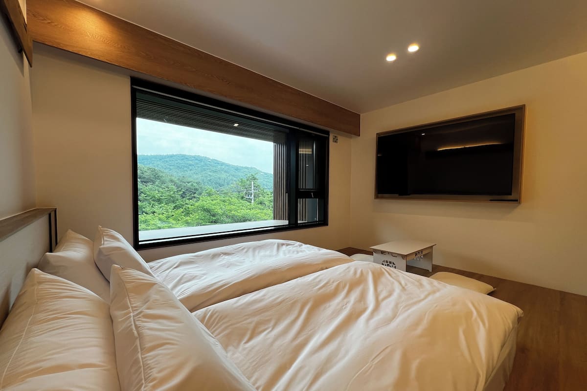 Ein Schlafzimmer mit zwei Betten, einem großen Fenster mit Blick ins Grüne, einem flachbildschirmfernseher an der Wand und einem kleinen Nachttisch.