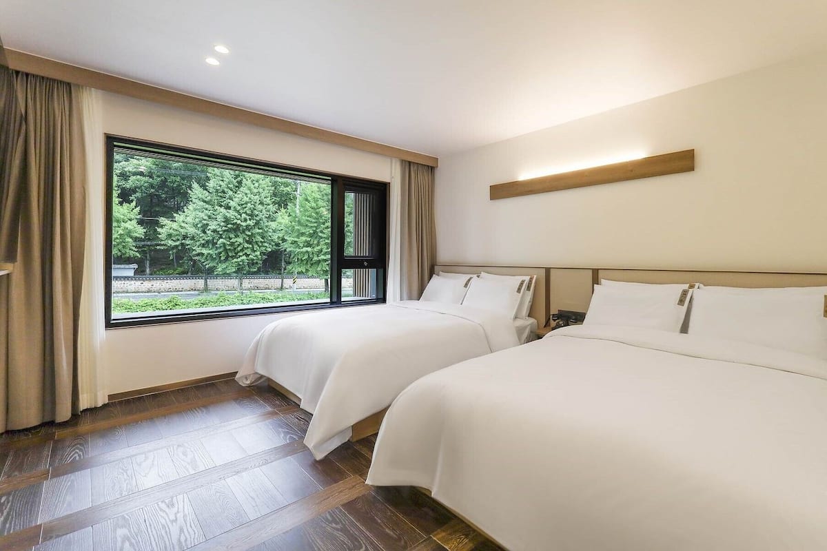 Ein Hotelzimmer mit zwei Betten, einem großen Fenster mit Blick auf Grünflächen, Holzböden und einem schlichten, klaren Design.