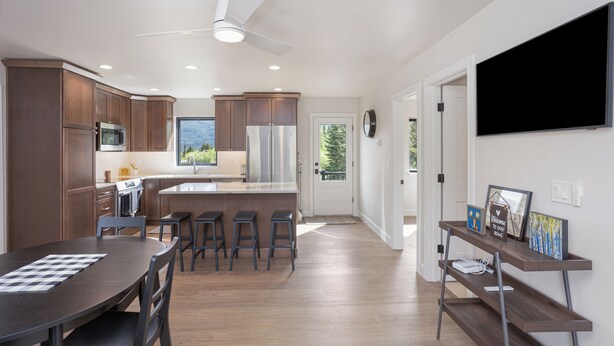 A modern kitchen with wooden cabinets, a central island with stools, a dining table, and a TV mounted on the wall. - Cozy Guest House In The Chugach Mountains  (Anchorage)