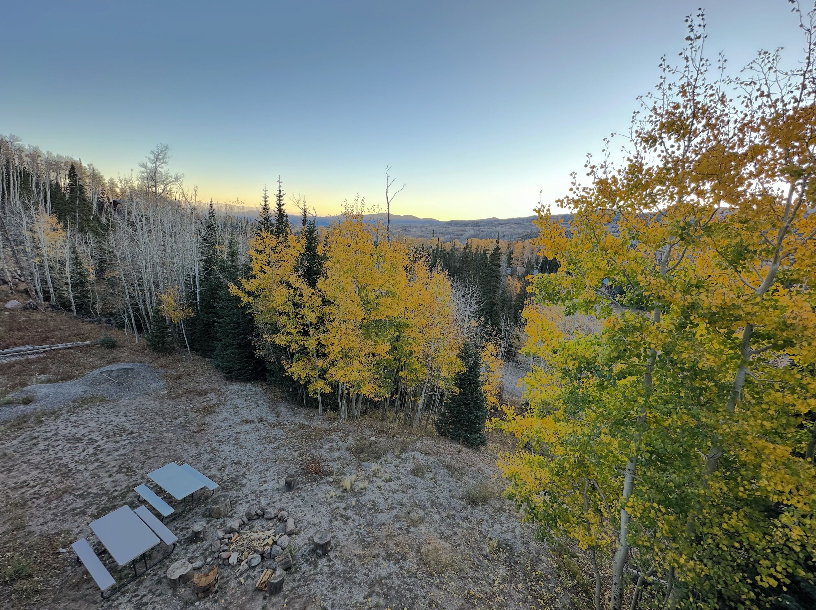 A campsite with a fire pit and metal tables, surrounded by trees with yellow leaves.