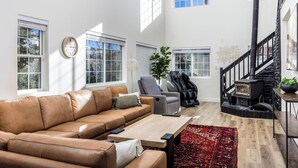 Een ruime woonkamer met een leren bank, een houten salontafel en een roodgekleurd tapijt. Er is een zwart haardvuur met een stenen omlijsting, een grijze fauteuil en een groot raam dat zorgt voor natuurlijk licht.