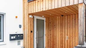 Eine Holzausfachung mit Glastür, ein Metallmülleimer und eine kleine Sitzgelegenheit im Freien.