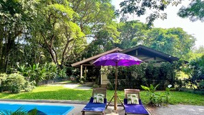 Un espace aménagé au bord de la piscine, avec deux chaises longues et un parasol violet, entouré d’une végétation luxuriante et avec un bâtiment visible au loin.