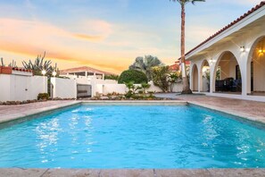 A swimming pool with a white wall and a covered patio area with arches and outdoor seating.