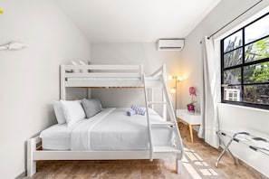 A bunk bed room with a white color scheme, a small bedside table with a lamp, and a window with a view of trees.