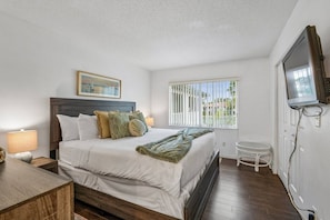 A bedroom with a bed, bedside lamps, a window with blinds, a small stool, and a television mounted on the wall.