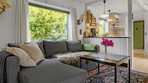 A modern living room with a grey sectional sofa, a glass coffee table, and a vibrant rug. A green door leads to the kitchen, which features white cabinetry and a wooden shelf. - 2 bedroom amazing home in Stege (Stege)