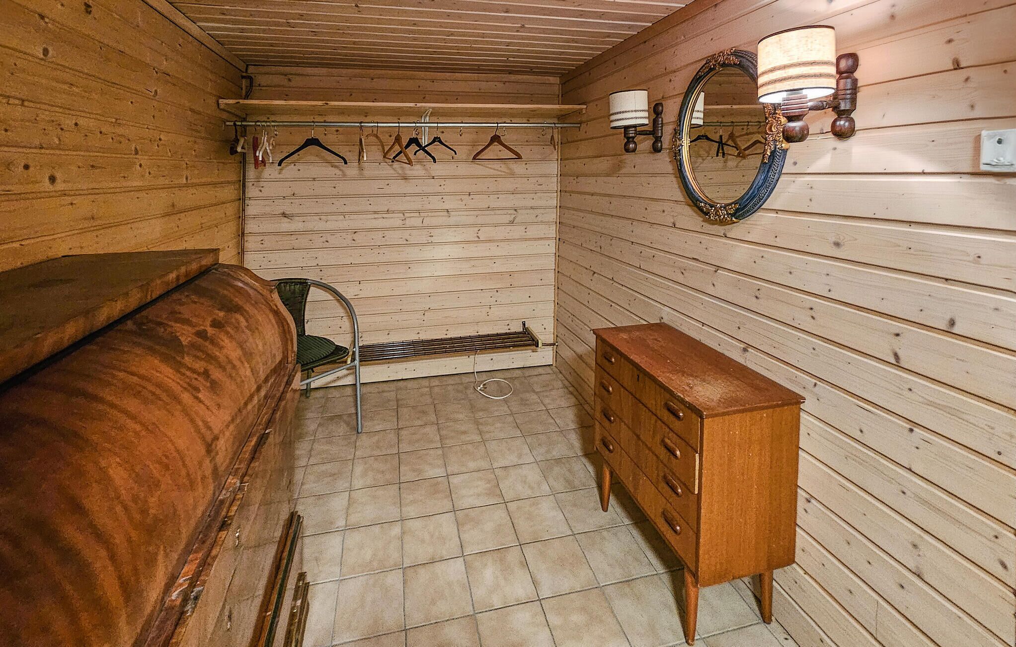 A wooden room with a bed, a dresser, a chair, a mirror, and a wall-mounted lamp.