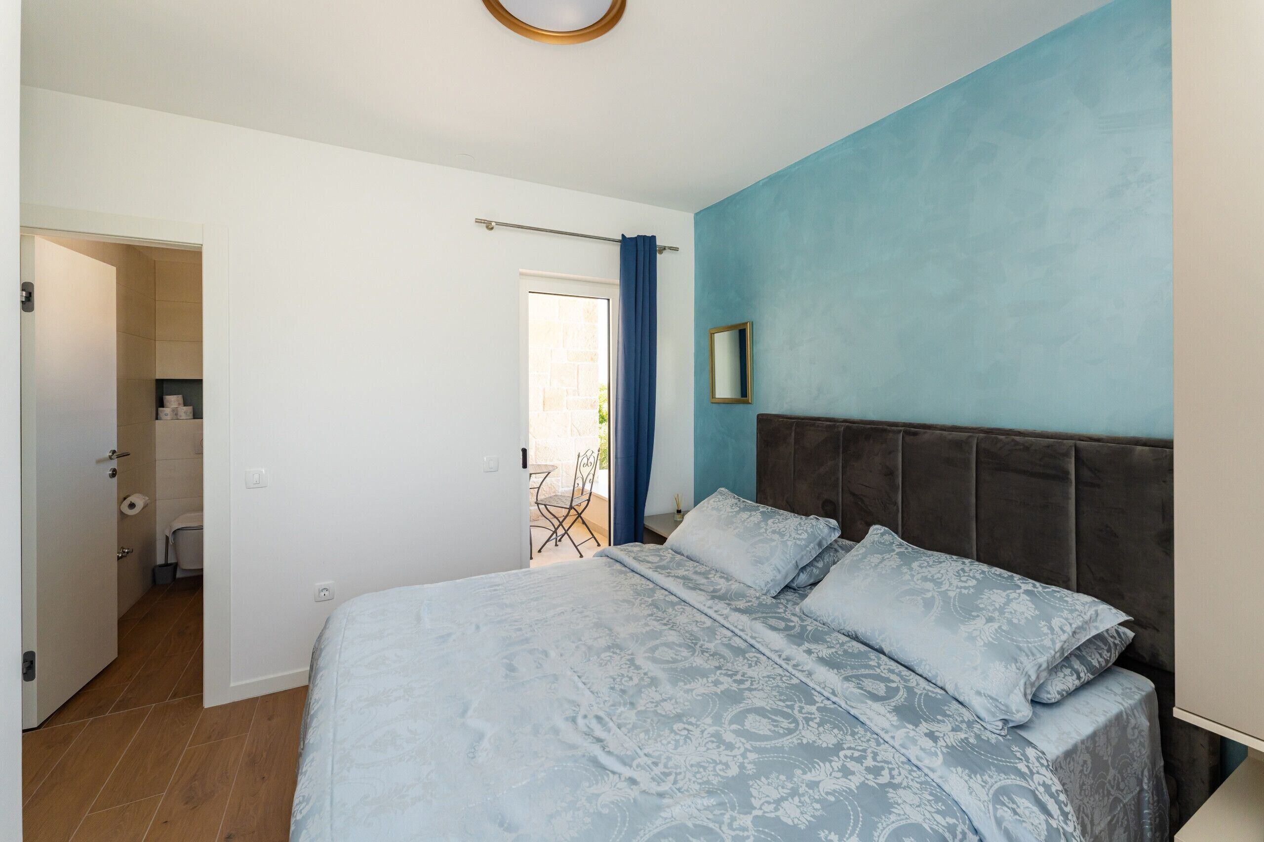 A bedroom with a bed, a headboard, a window with curtains, a door, and a small table with chairs.