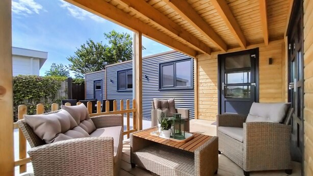 Een overdekt terras met rieten meubels en een tafel, met uitzicht op een modern huis met een blauwe gevel.