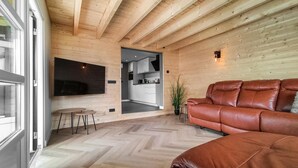 Een modern interieur met een houten plafond, een flatscreen televisie aan de muur bevestigd, een leren bank en een keukenruimte zichtbaar door een open deur.