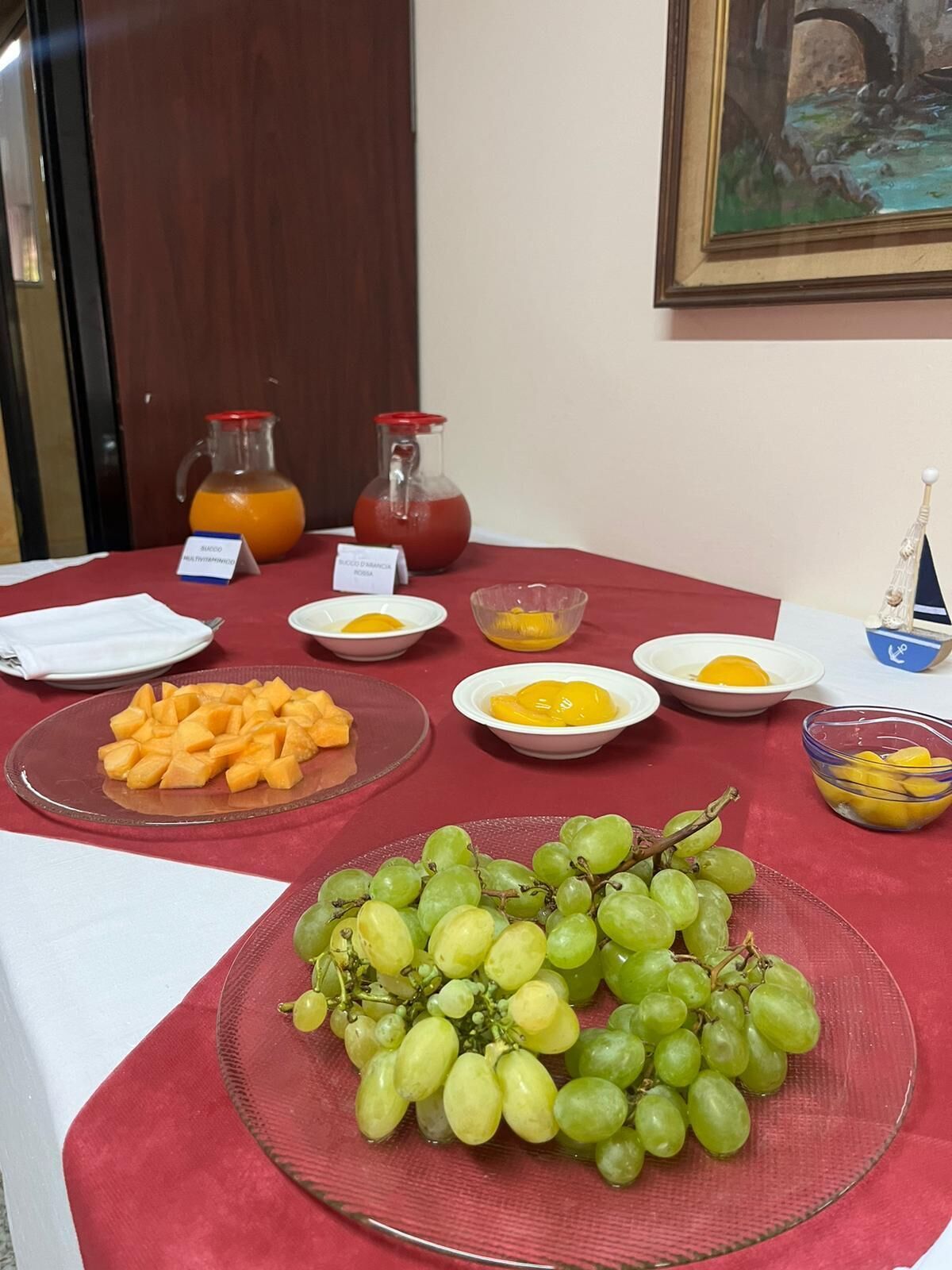 A breakfast buffet table with various fruits, including grapes and mango chunks, and two pitchers of orange and tomato juice.