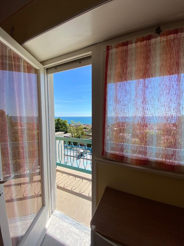A room with a sliding glass door leading to an outdoor balcony with a view of the sea and buildings.
